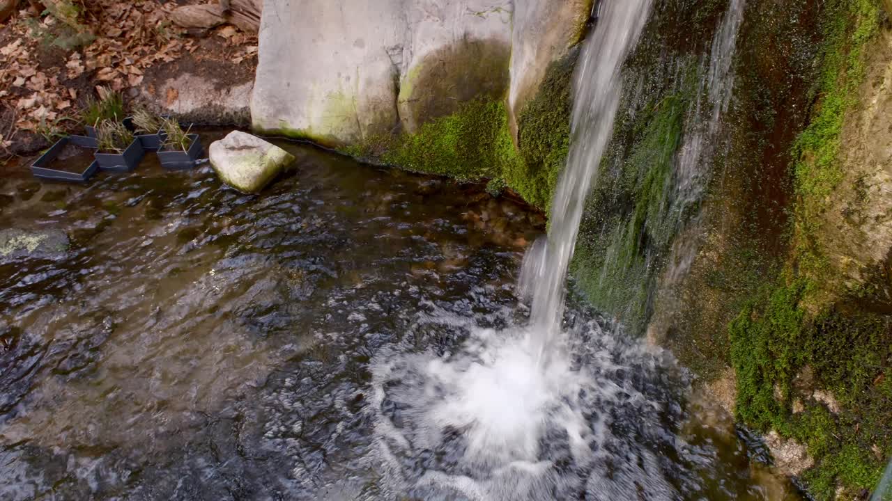 A small waterfall cascades into a rocky pool, creating ripples and splashes in the clear water below. The scene is surrounded by moss-covered rocks, evoking a serene natural setting.