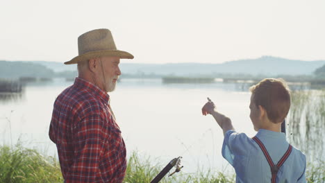 vista trasera de un abuelo y un nieto sosteniendo sus cañas de pescar y hablando en la orilla del río