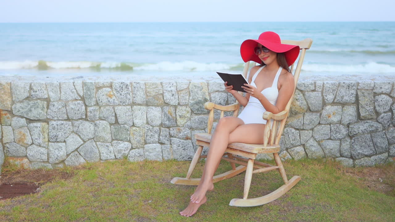 Sitting in a rocking chair with the surf rolling in the background, a young woman in a bathing suit and floppy sun hat reads a book,
