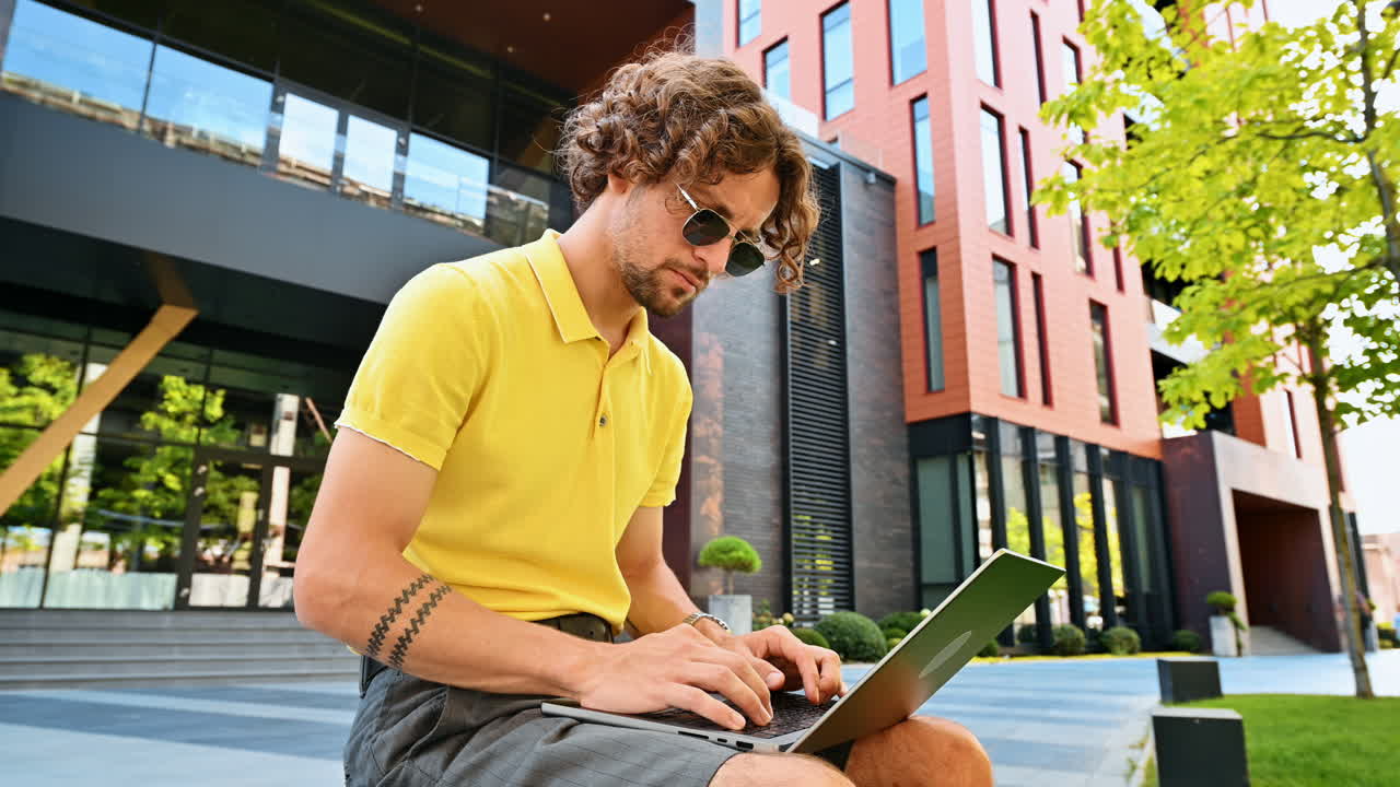 Uomo con camicia gialla che parla stando in piedi su una panchina e lavorando su un laptop
