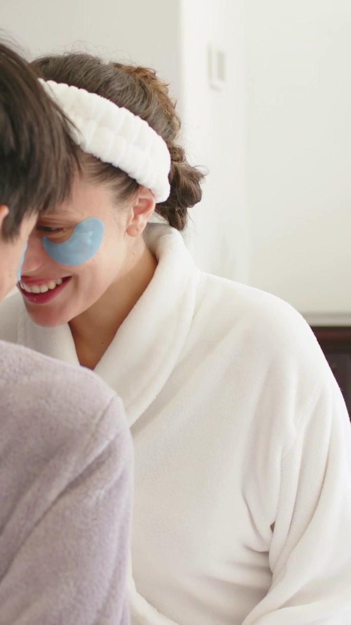 Vertical video: Smiling woman in robe leaning, chatting as man entering at vanity with blue patches