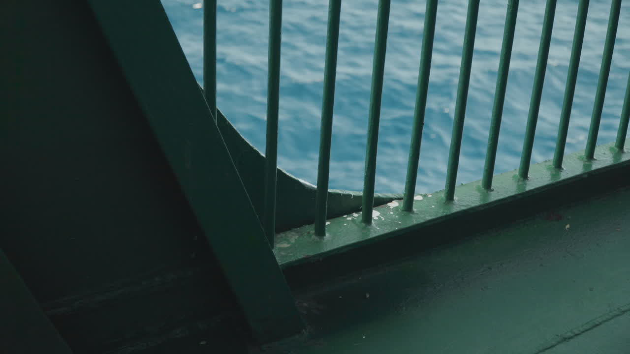 Nahaufnahme: Fährgeländer vor tiefblauem Wasser, abgenutzte Farbe und Nieten, schattiger Decksrand mit Salzwasser-Gischt. Raue, industrielle Stimmung, detailreich mit filmischem Korn für redaktionelle und kommerzielle Zwecke.