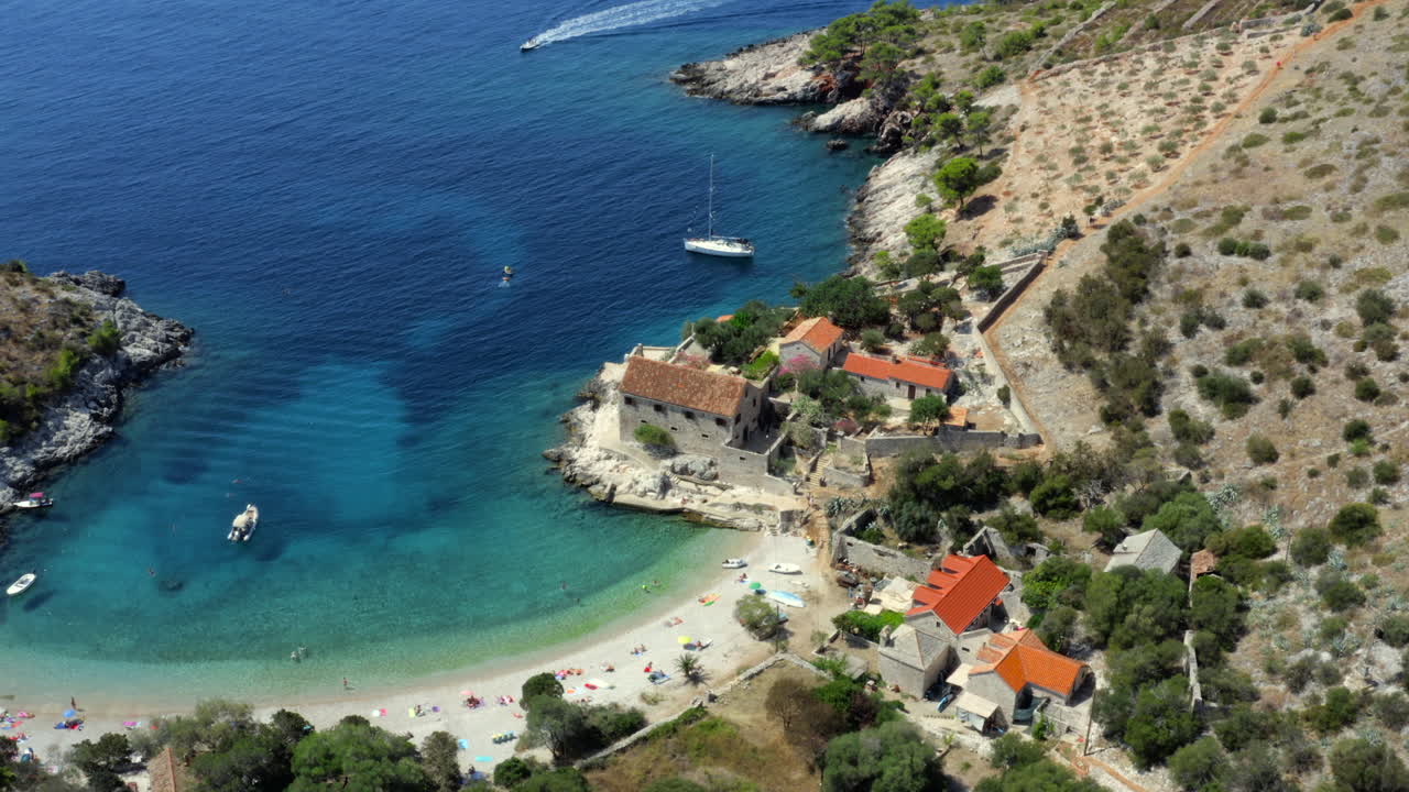 Aerial establishing shot of secluded Mediterranean beach with turquoise water, boats and stone houses with red roofs on a sunny day. Hvar, Croatia