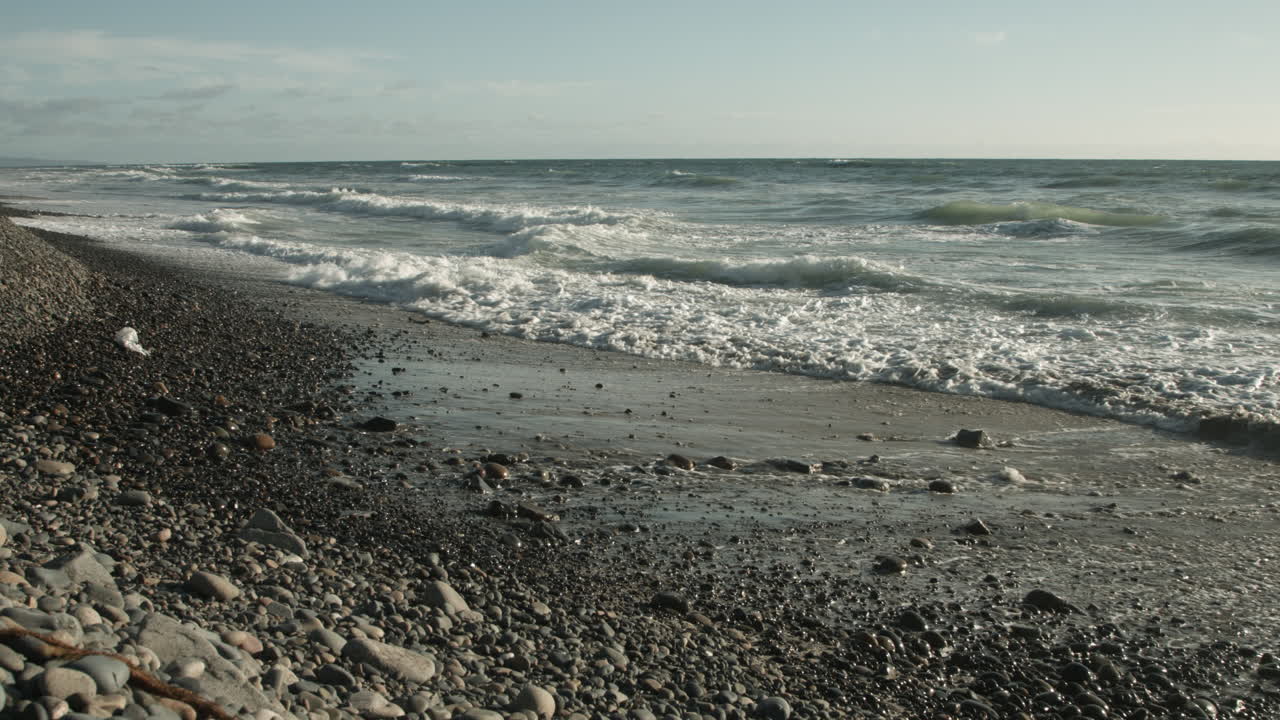 Gentle ocean waves roll onto a tranquil beach scattered with sand and pebbles, creating a soothing, natural rhythm under soft coastal light—perfect for themes of peace, nature, and reflection.