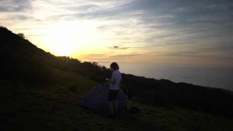 young man starts flying drone at sunset with ocean view at campsite