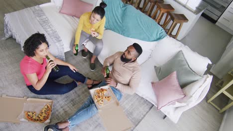 ángulo alto de diversos amigos felices femeninos y masculinos disfrutando de cerveza y pizza en casa, cámara lenta