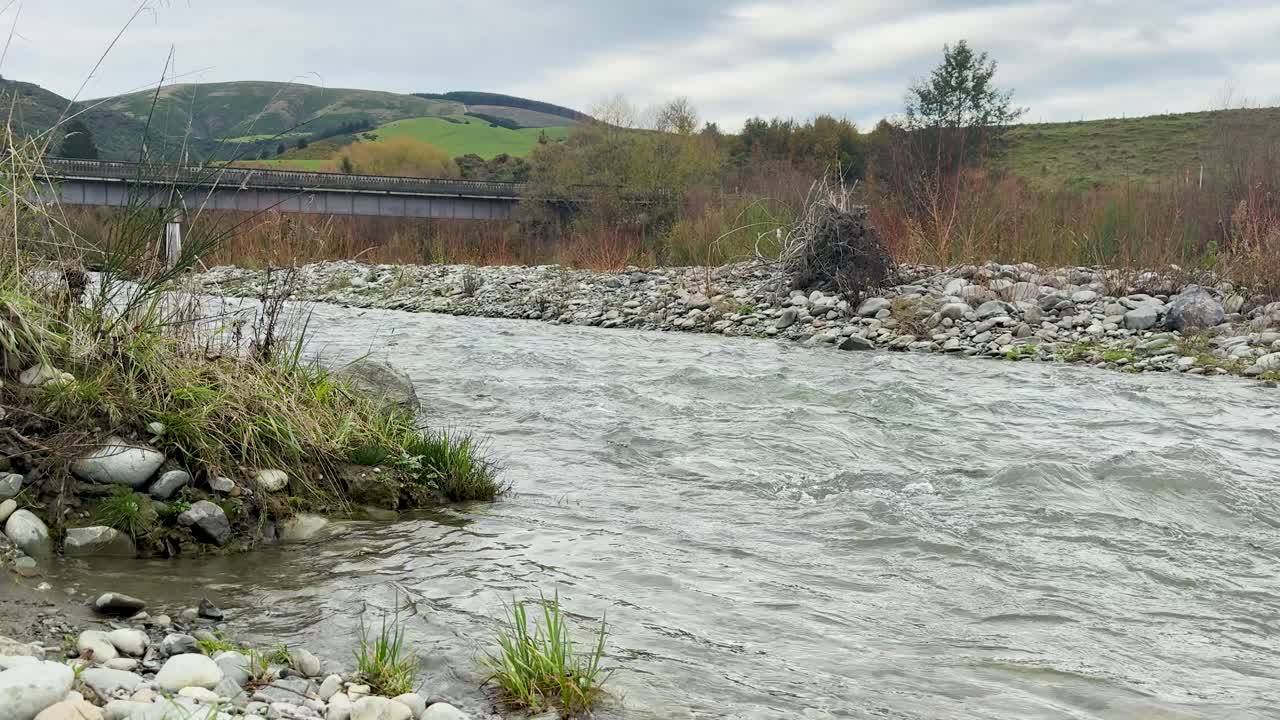 A steady current moves through a rocky, grassy stream bordered by reeds and wildflowers under overcast daylight, with distant hills and a rustic bridge