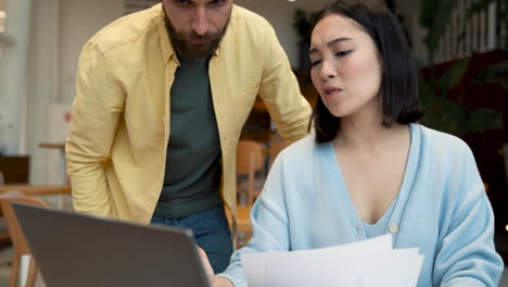 Una-Mujer-De-Negocios-Que-Muestra-Documentos-En-Papel-Y-En-Su-Computadora-Portátil-A-Un-Compañero-En-Una-Reunión-En-Una-Cafetería-1