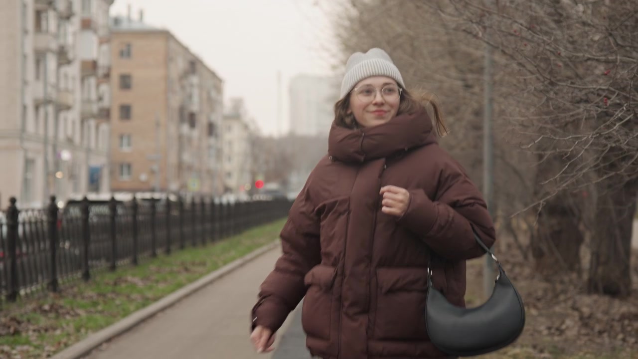 Woman confidently walking in city background. Smiling lady with glasses in urban scene during daylight. Closeup of cheerful woman in warm coat walking confidently with blurred city behind