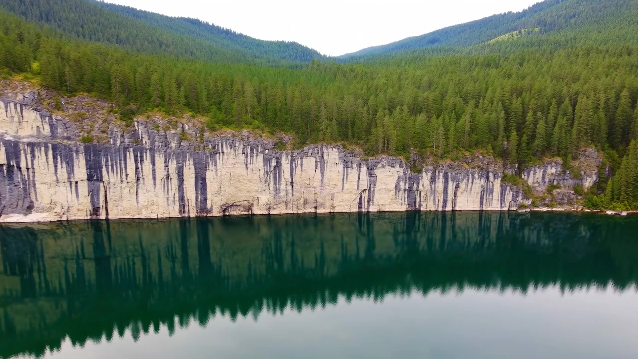 Vidt luftfoto av en rolig turkis innsjø avgrenset av steinete klipper, tett eviggrønn skog og fjerne grønne fjell, med stille vann som reflekterer strandlinjen i mykt dagslys.