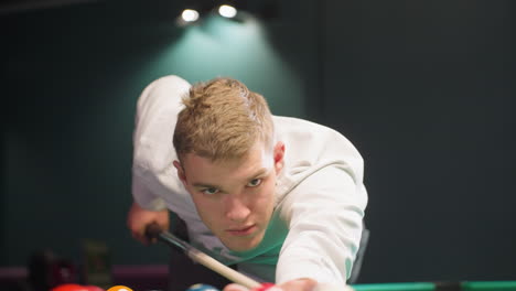 close-up front view of young man in white shirt leaning over pool table in dark billiard room, gripping cue stick with precision. focused expression as he lines up strategic