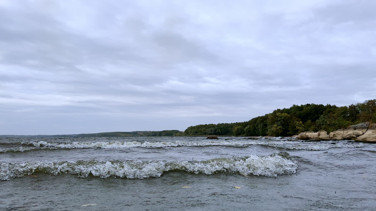 Quick waves approaching the shore of the lake. Sky above the water is covered with grey cloudscape. Gloomy summer day footage.