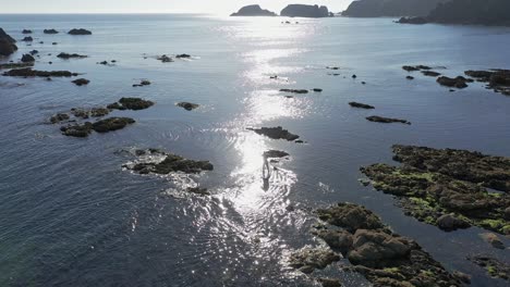 Vista-Aérea-Del-Surf-De-Remo-En-Aguas-Tranquilas-Con-El-Sol-Reflejándose-A-Lo-Largo-De-La-Costa-Rocosa-En-El-Sur-De-Irlanda-En-Un-Día-Tranquilo