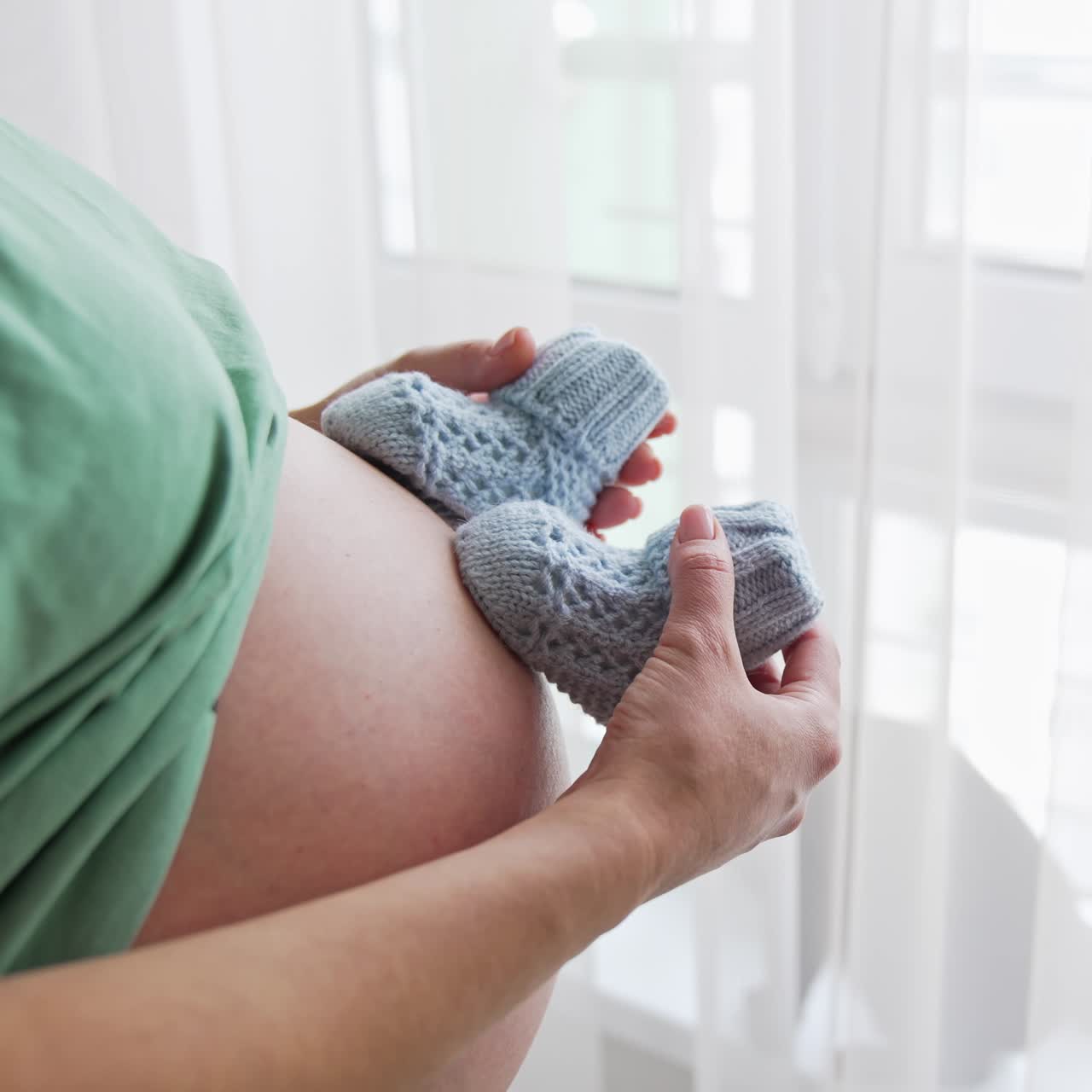 Pregnant lady stands with her face to the window. Mother plays with baby socks on her bag belly. The cat walks by on the window-sill behind the curtains