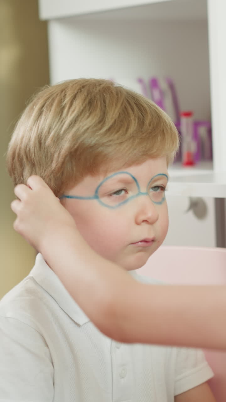 la niña pinta el rostro del hermano menor con lápiz de color en cámara lenta. la hermana arregla el cabello cerca de la oreja del hermano dibujando gafas en primer plano. los hermanos se divierten juntos