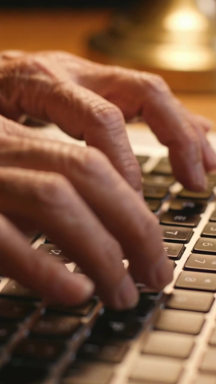 Vertical video: Lowering onto keys, senior hands tapping laptop keyboard at wooden desk, lamp lit