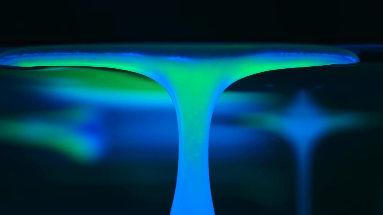 Scena in movimento ravvicinata che mostra un liquido lucido tirato verso il basso da una sottile membrana superiore in una colonna verticale continua e un sottile flusso, con un bagliore ciano neon, smeraldo e verde acqua su uno sfondo scuro.