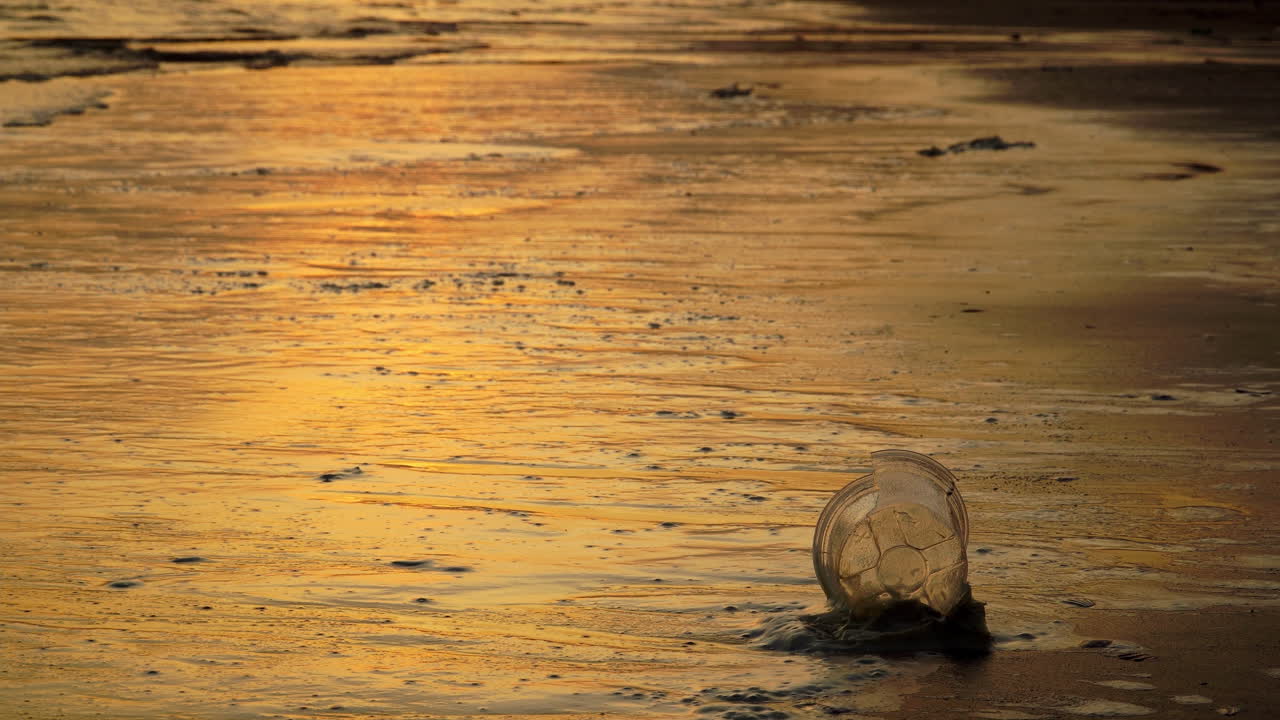vaso desechable de plástico en la playa de arena mientras las olas del océano golpean la costa, la puesta de sol de la hora dorada