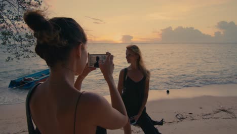 two women photographing each other on a beach sunset