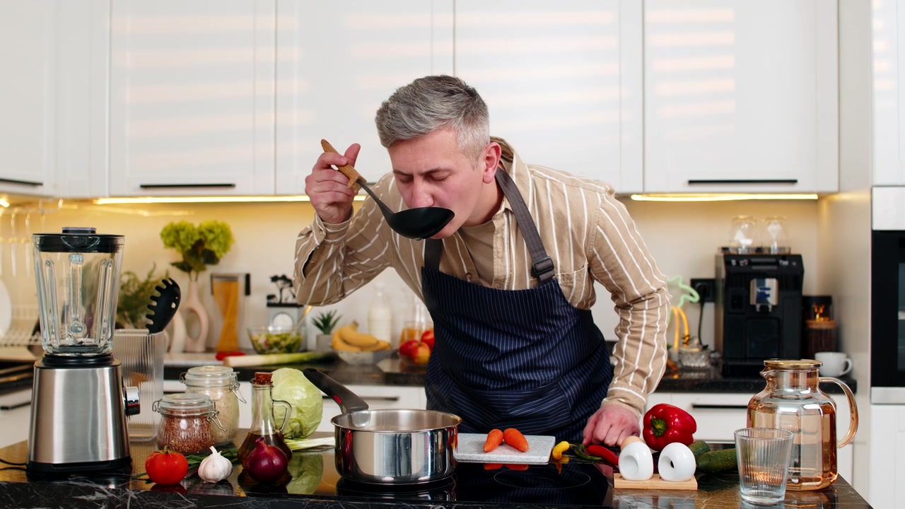Mann mittleren Alters kocht frische Diätsuppe in der Küche, riecht das Aroma und probiert die gesunde hausgemachte Mahlzeit.