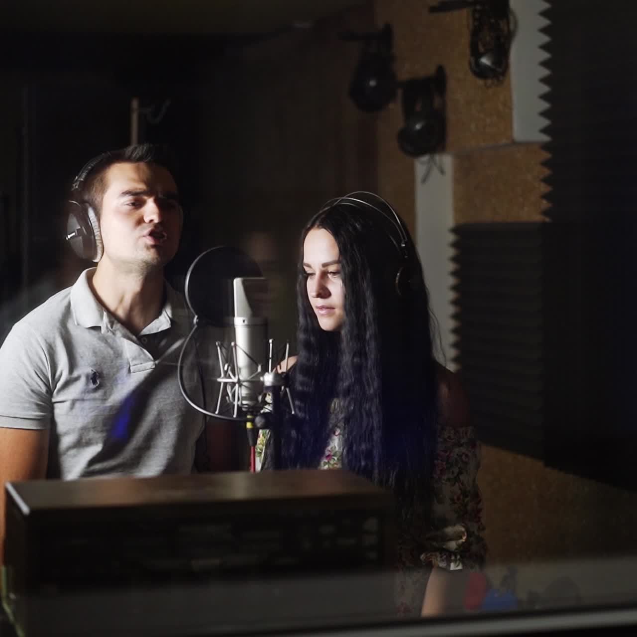 Male and female duet performing song in the studio. A couple wearing headphones sing in front professional microphone