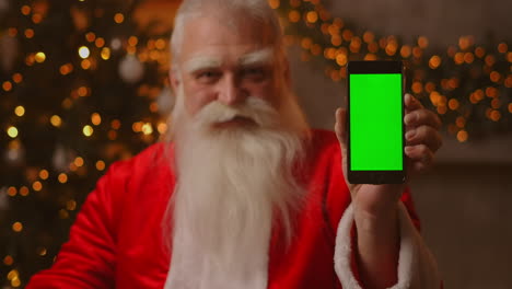 Santa-Claus-is-sitting-on-the-sofa-in-the-background-of-a-Christmas-tree-and-garlands-holding-a-mobile-phone-with-a-green-screen-pointing-at-it-with-his-finger.-Santa-holds-a-phone-with-a-chromakey-on-the-screen