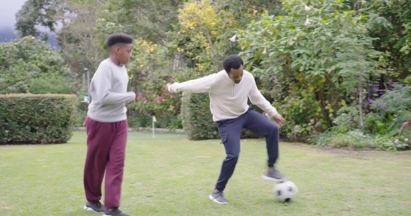 Pria dewasa dan remaja Afrika menggiring bola sepak di lapangan rumput, dewasa menunjukkan dan mengajar, remaja meniru