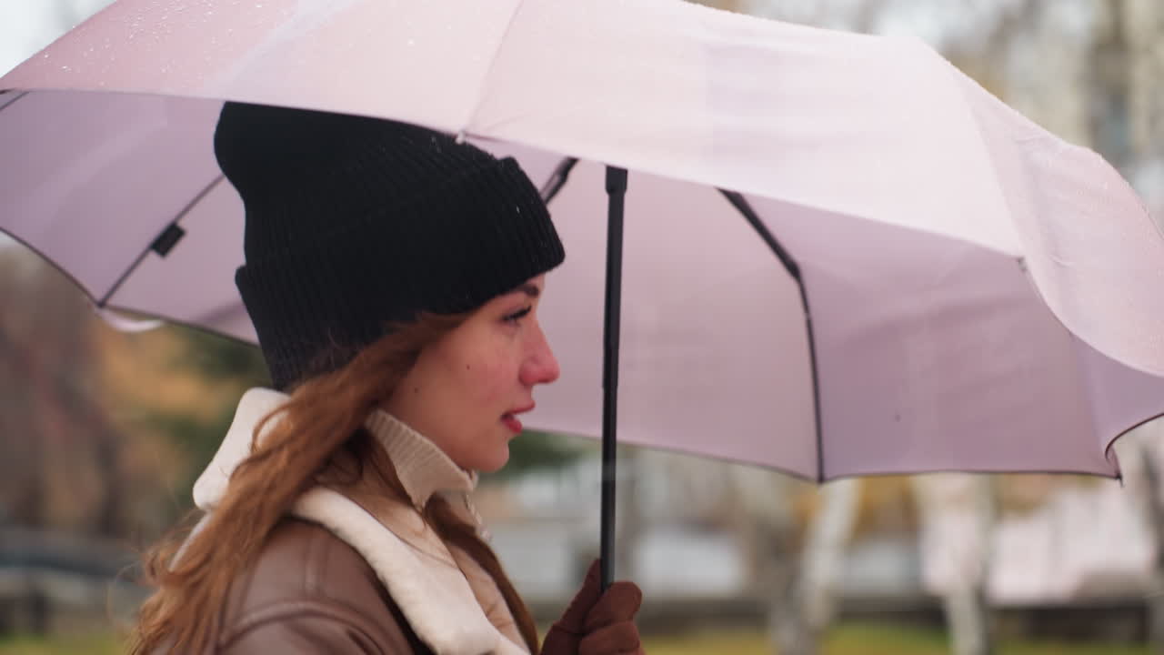 Close shot of cheerful girl holding umbrella as wind blows gently with light snowfall running outdoors wearing black knit cap and brown shearling jacket strands of hair flowing in soft winter breeze