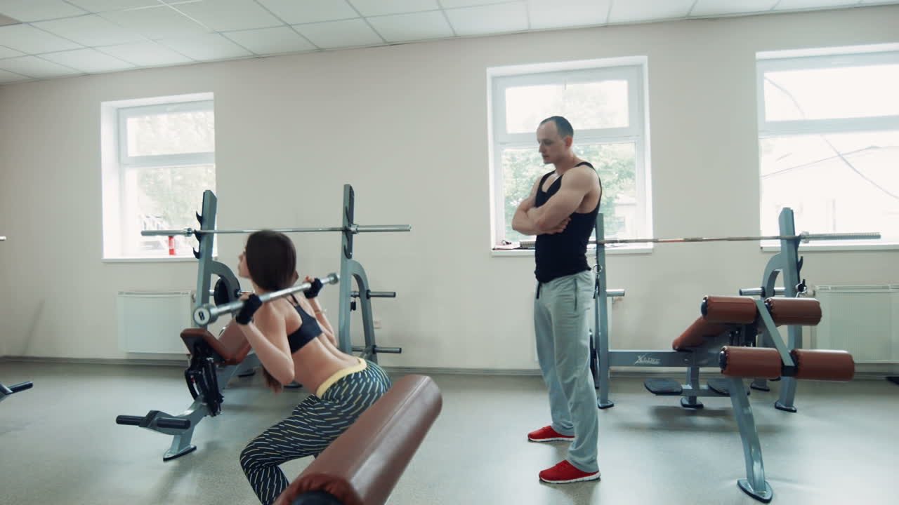 Woman doing squats with barbell, supervised by a trainer in the gym.