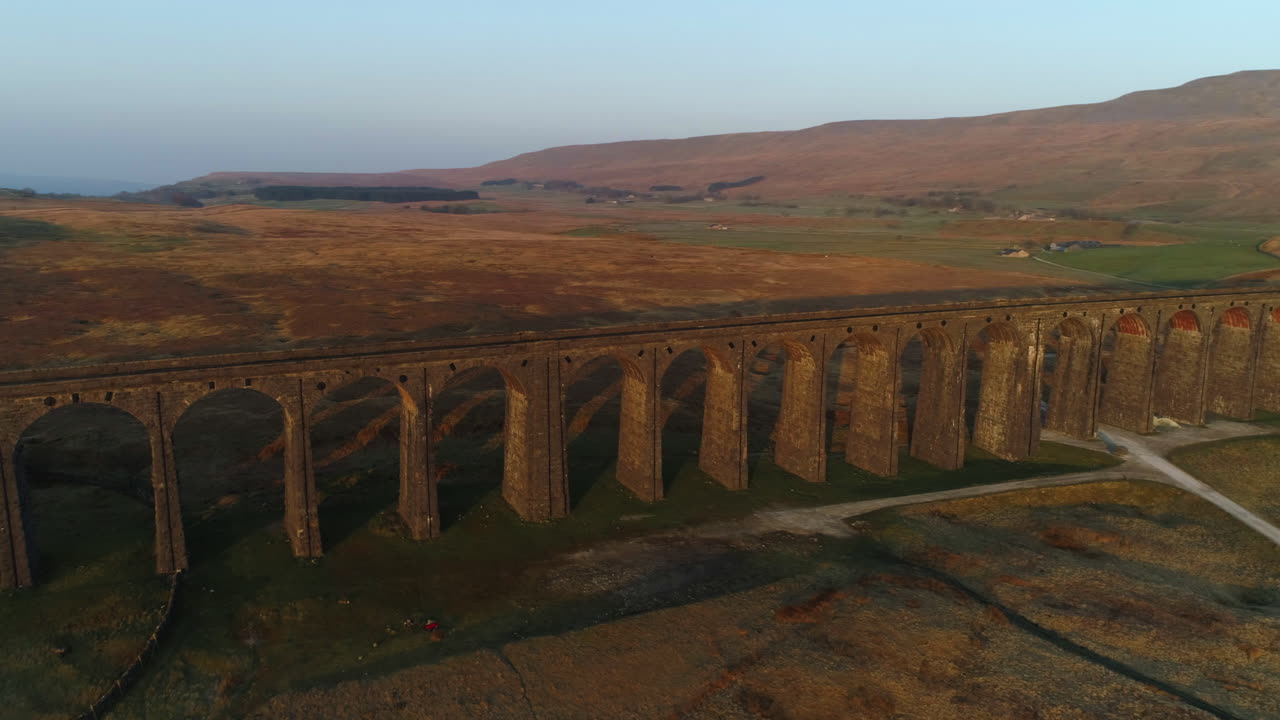 Ripresa aerea con drone che ruota intorno al ponte ferroviario del Viadotto di Ribblehead durante una splendida alba estiva nello Yorkshire Dales, Inghilterra, Regno Unito, con le 3 Cime del Monte Whernside sullo sfondo.