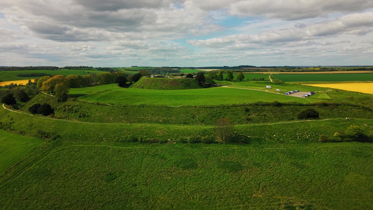 Wide aerial drone view of Old Sarum, the ancient Iron Age hillfort and historic earthwork site near Salisbury in Wiltshire, surrounded by green English countryside, open farmland and dramatic cloudy s