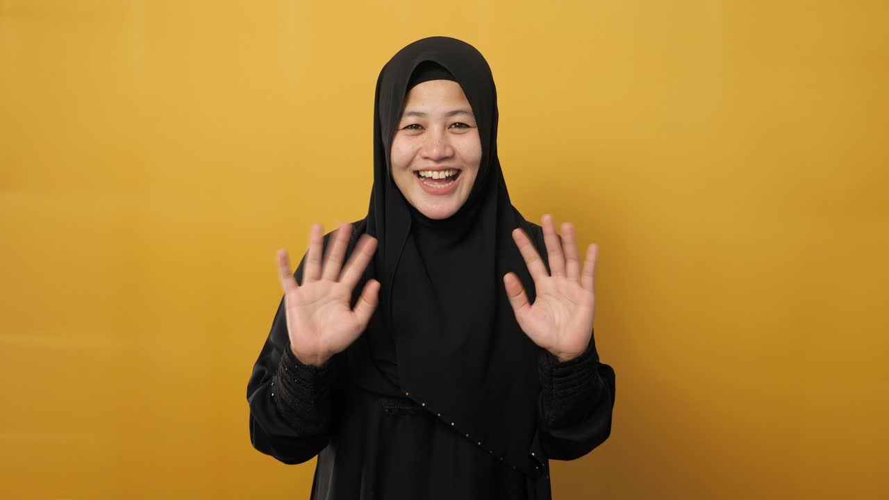 히자브를 입은 무슬림 여성이 카메라를 바라보며 미소 짓고 손을 흔들며 안하세요 또는 작별 인사 제스처를 노란색 배경에