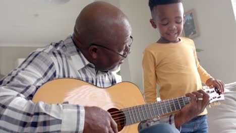 happy african american grandfather and grandson playing guitar together at home, slow motion