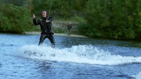 Glücklicher-Wakeskifahrer,-Der-Während-Der-Bewegung-Auf-Dem-See-Hinter-Dem-Boot-Mit-Den-Händen-Zeichen-Macht