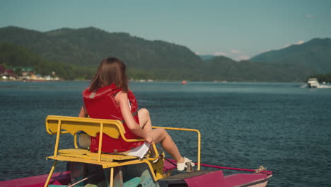 young woman in red life jacket sits in catamaran looking at boat. tourist enjoys landscape on clear lake slow motion backside view. summer fun on water in recreation area