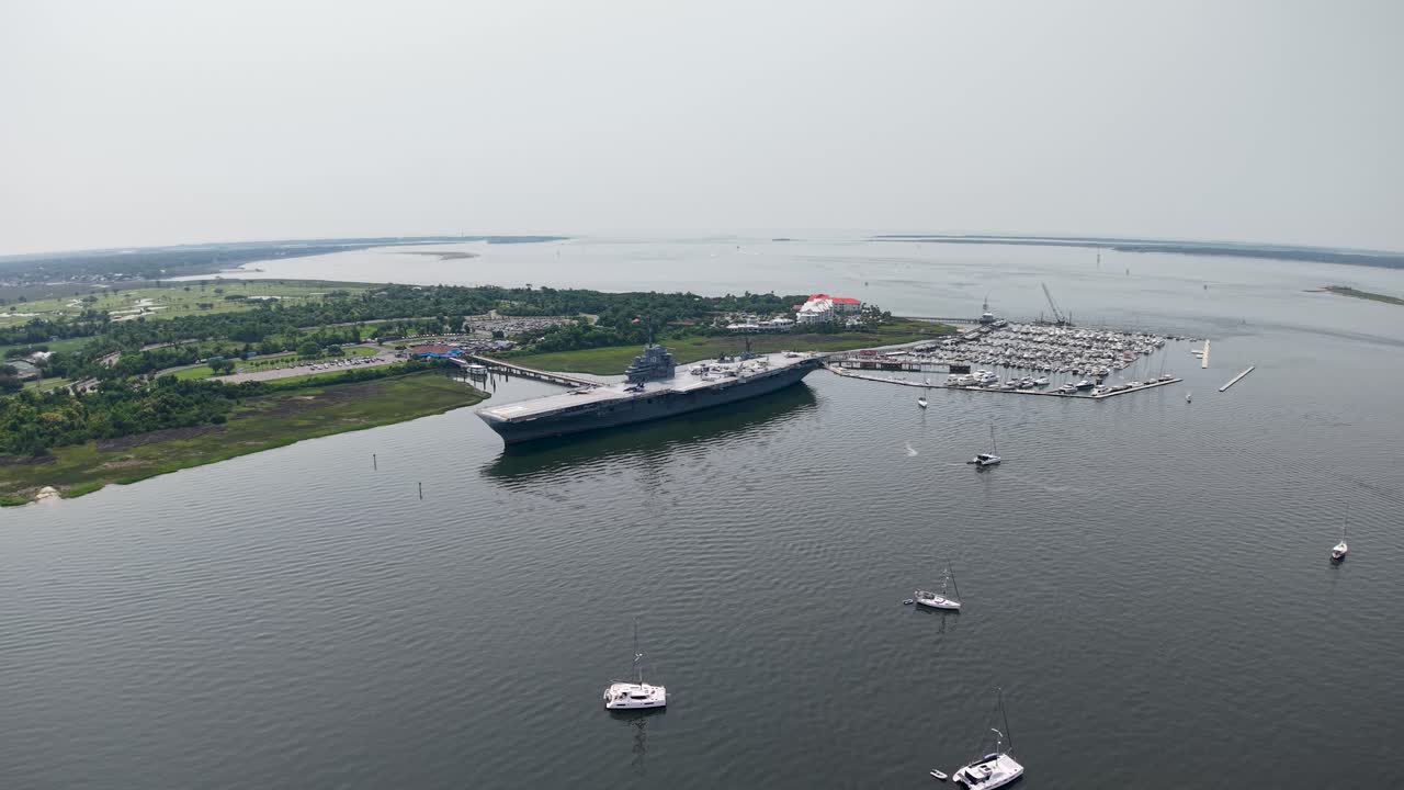 Drone video of the historic USS Yorktown aircraft carrier anchored in Charleston Harbor, with marshy wetlands, boats and a sprawling waterfront landscape under a clear sky