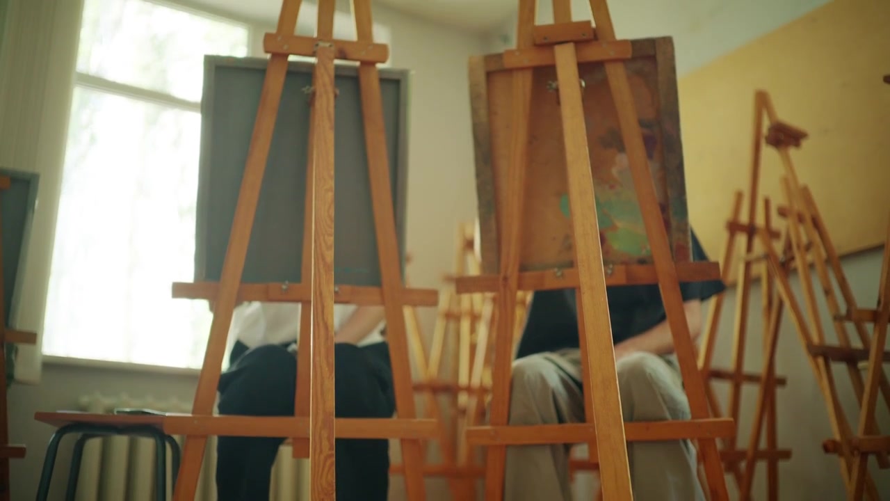 Cavalletti di legno disposti all'interno di una luminosa aula d'arte, che rappresentano l'educazione creativa, i laboratori artistici e gli spazi di apprendimento dedicati alle arti visive e al design.