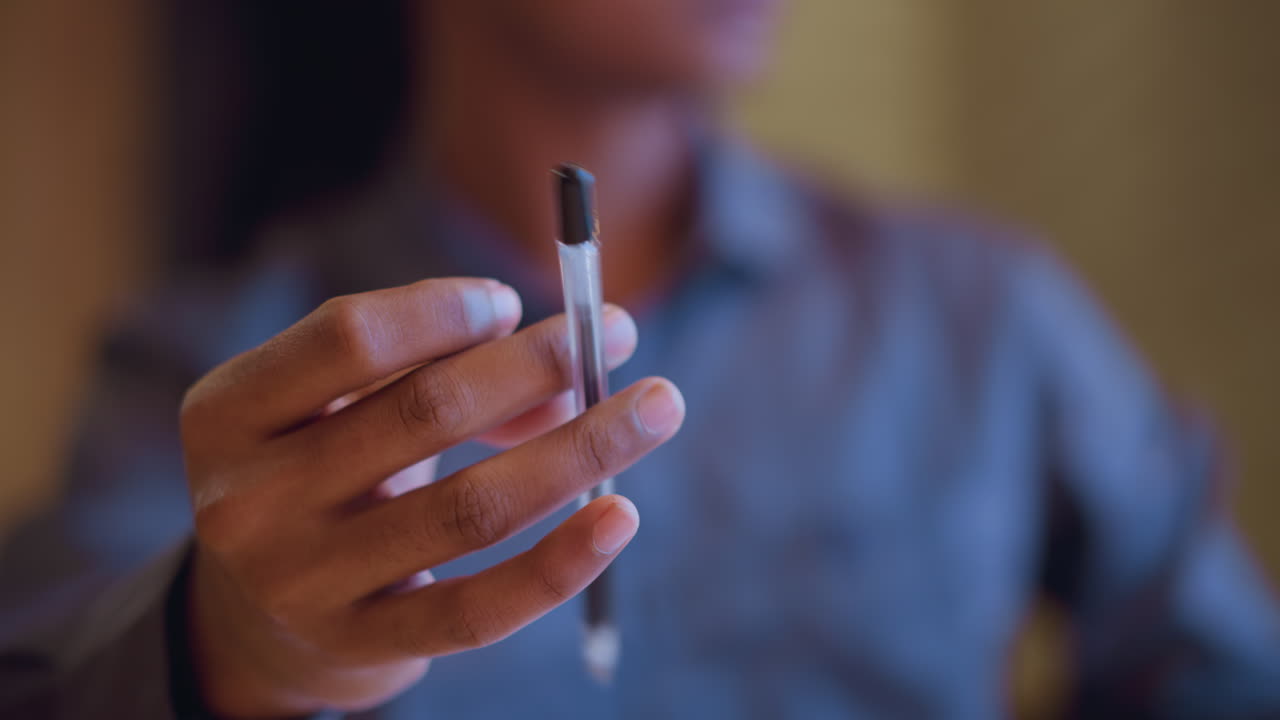 Close-up of man holding pen upright between fingers in professional setting, suggesting pause before speaking, readiness to write, or expression of idea during thoughtful moment in office environment