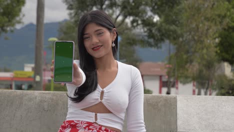 Cinematic-close-up-clip-of-a-young-brunette-latin-girl-using-her-smartphone-and-showing-the-screen-that-has-a-green-screen