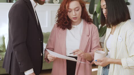 young redhead woman showing paper report to her two multiethnic colleagues and debating together 1