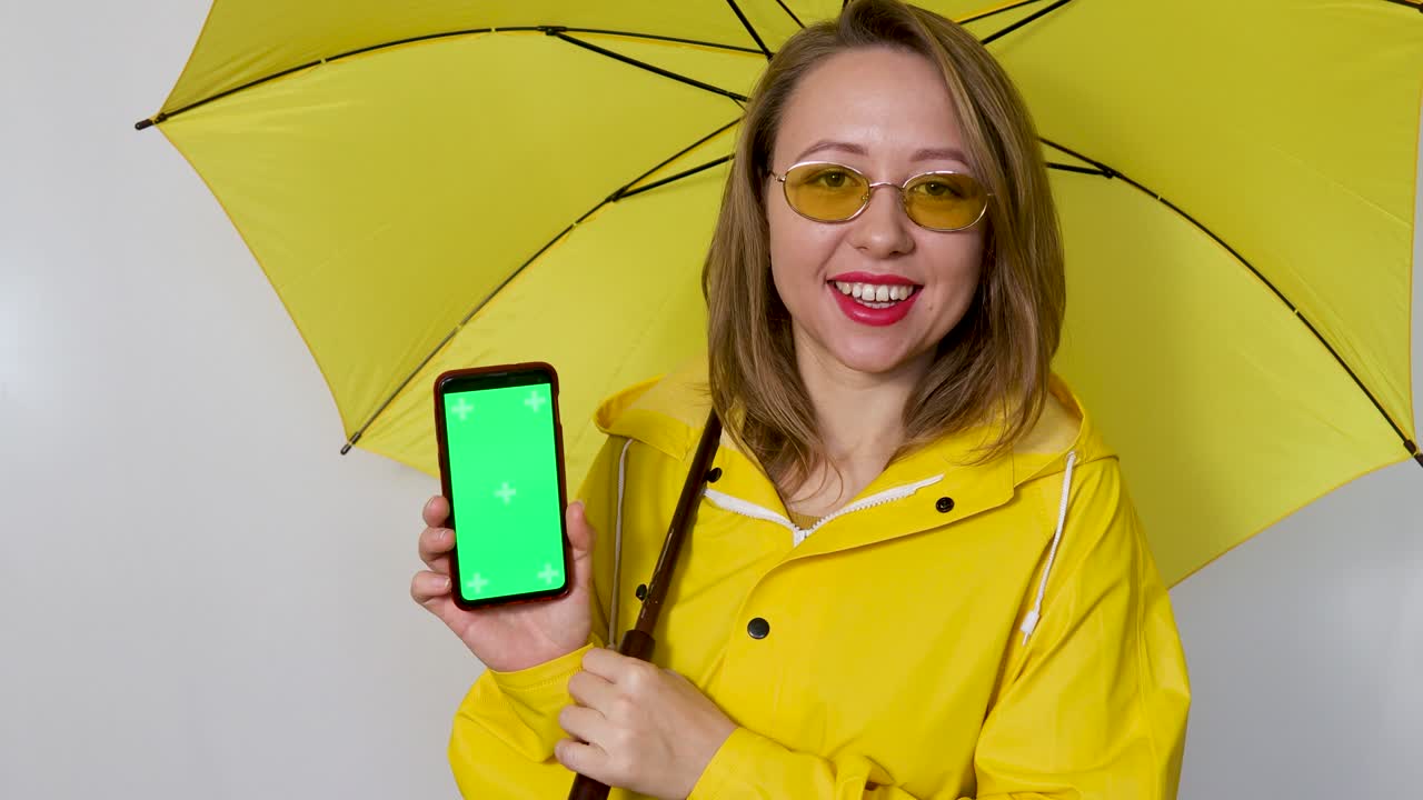 노란색 레인코트를 입고 우산을 쓴 소녀가 시청자에게 녹색 화면을 보여줍니다. 색 배경에 고립되어 있습니다.