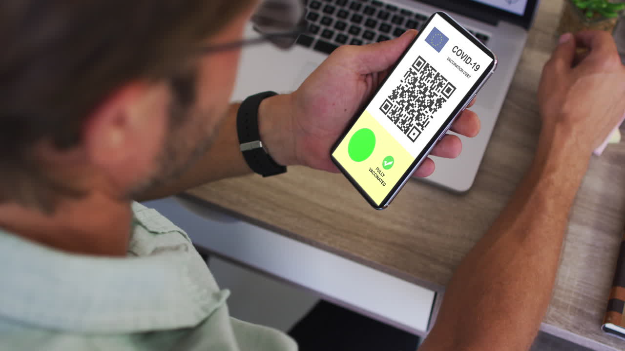 Man at desk holding smartphone with covid vaccination certificate, eu flag and qr code on screen