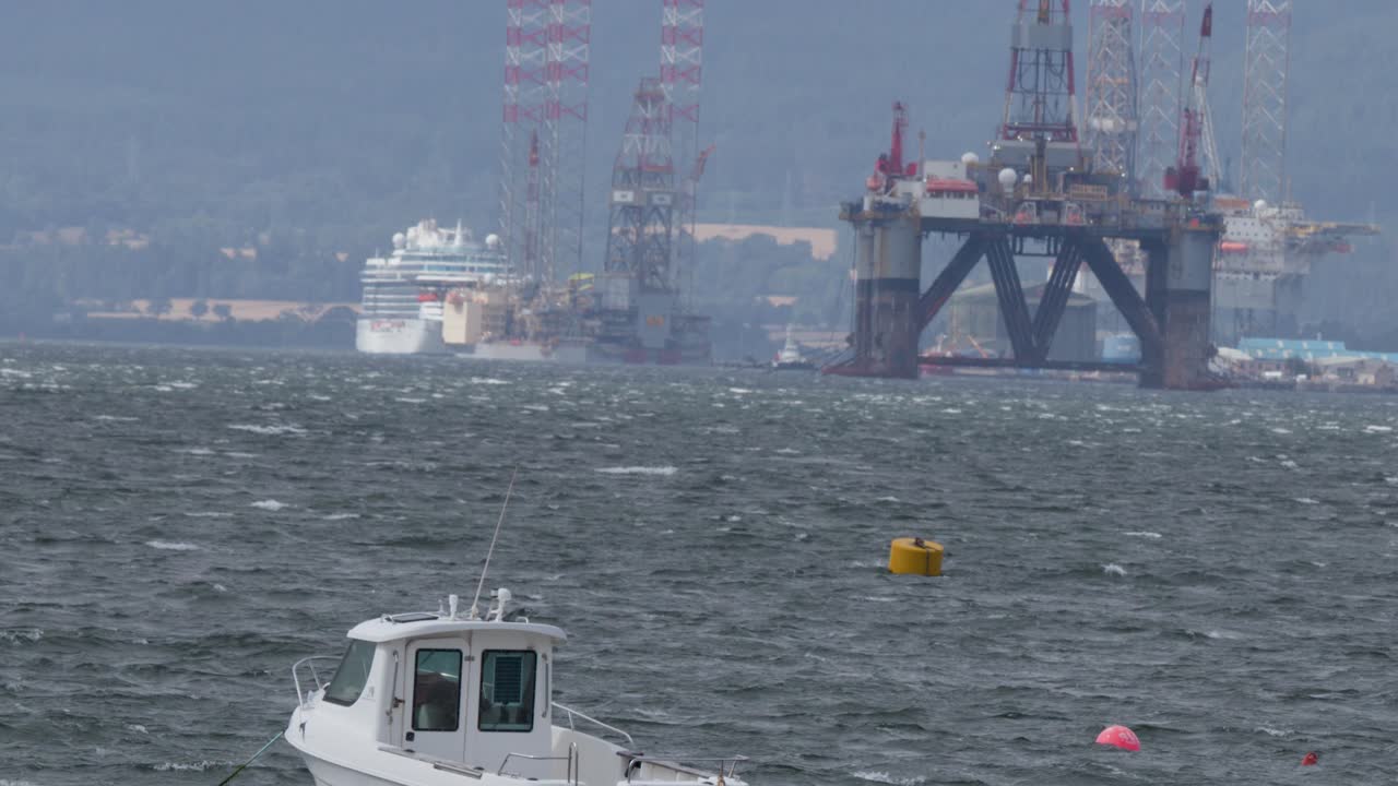 White boat drifts on choppy sea near industrial rigs, overcast daylight, distant mountains visible