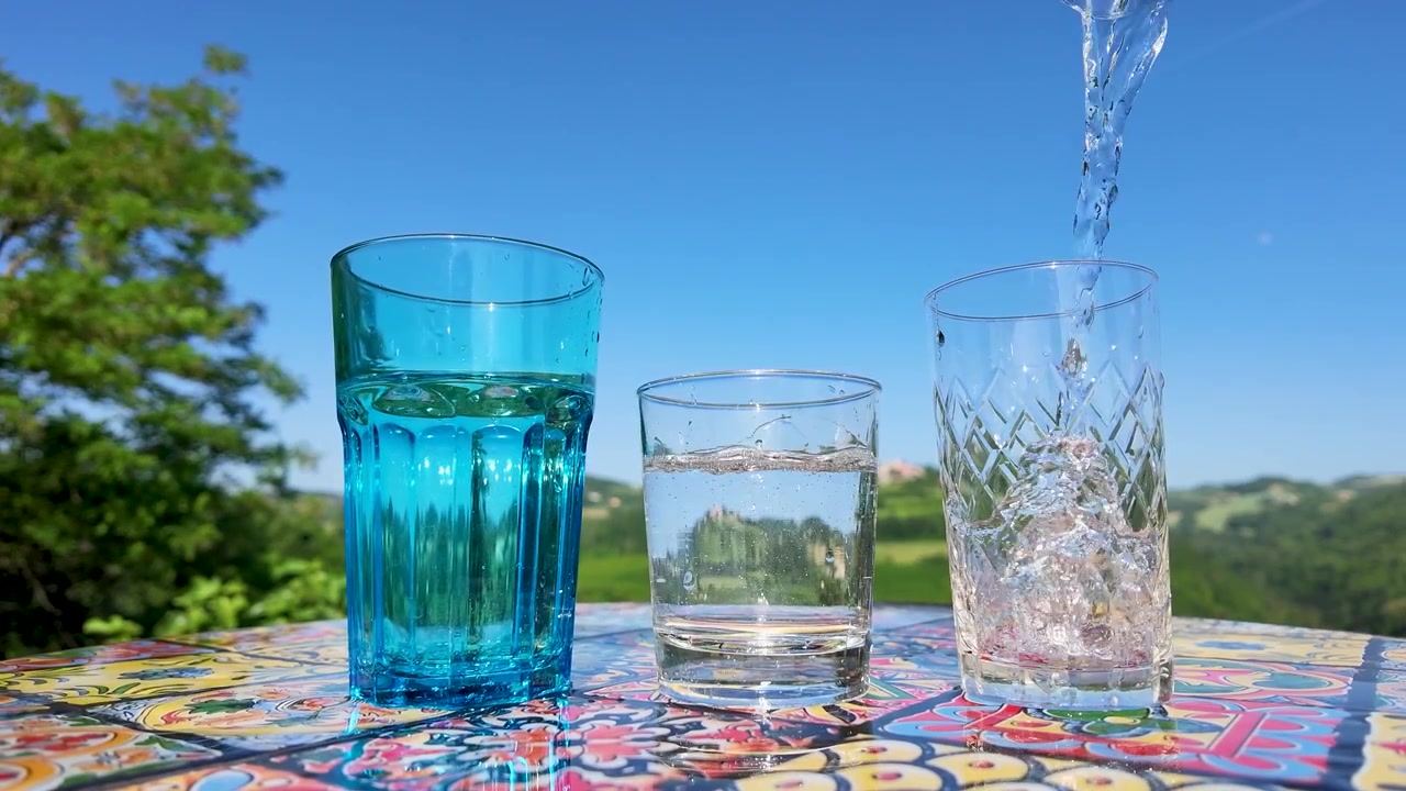 Acqua frizzante si versa in tre bicchieri su un tavolo colorato sotto un cielo azzurro brillante.