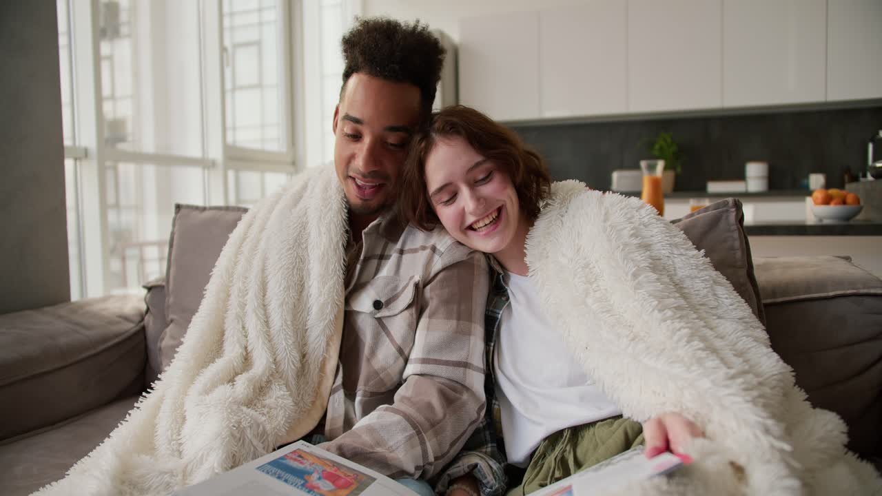 Happy young man with Black skin color in a plaid shirt sits with his girlfriend on a modern gray sofa wrapped in a white wool blanket. They read a book and stories in a modern apartment while spending time together