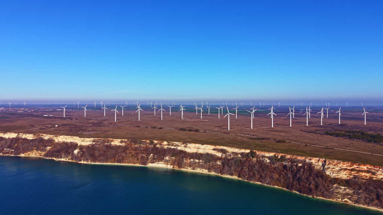 Wind turbines stand along a coastal landscape with clear skies and a blue sea in the distance during daytime