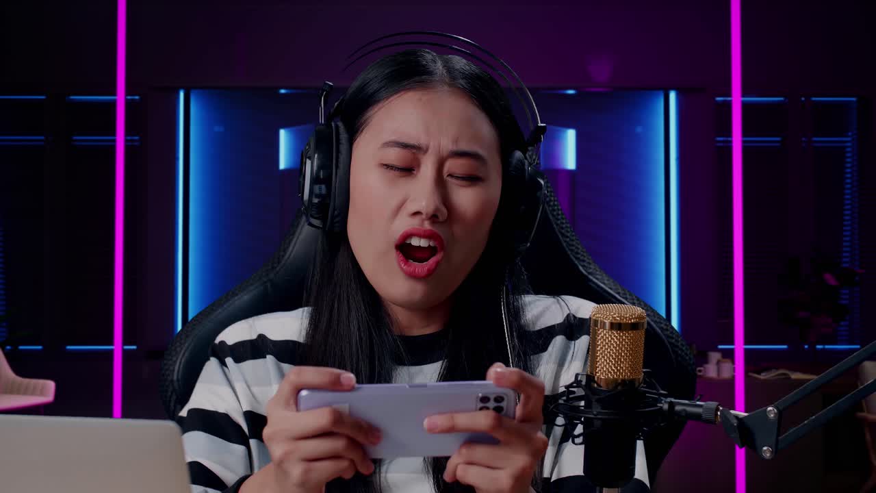 헤드폰과 컴퓨터를 가진 화가 난 아시아 여성 게이머가 방에서 네온 조명으로 조명된 휴대 전화 게임을 잃고 있습니다.