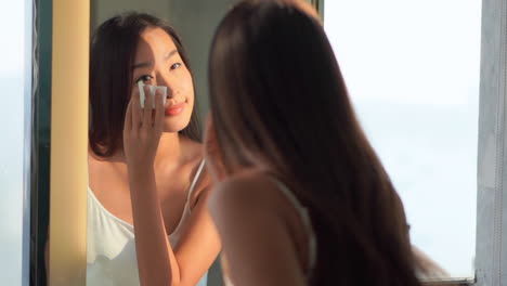 asian woman using cotton pads for cleansing the face of cosmetics with tonic or micellar cleansing water standing near the mirror