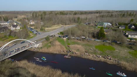 static aerial drone footage of rafts, kayaks and paddleboard boats rowing and paddling along a narrow brown colored river that flows under a bridge during a sunny day when spectators are on shoreline.