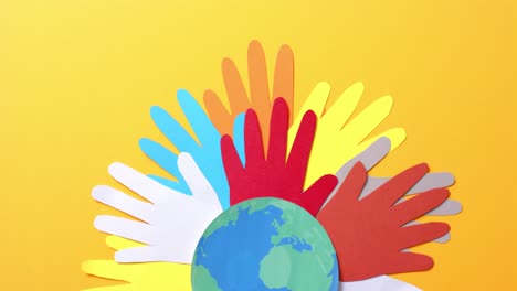 Close-up-of-hands-together-with-globe-made-of-colourful-paper-on-yellow-background-with-copy-space